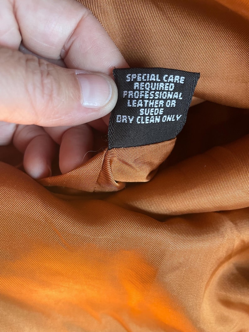 May include: Close-up of a brown leather jacket's care label. The label reads "Special care required professional leather or suede dry clean only."