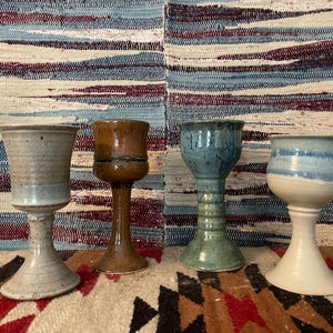 May include: Four ceramic goblets with distinct glazes: one speckled light gray, one glossy brown with a dark horizontal band, one mottled blue-green, and one with a cream base and a blue-dipped rim, all featuring flared stems and wide bases.