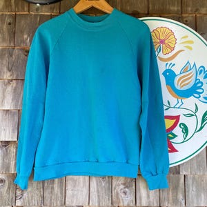 Op de afbeelding: Een turquoise crewneck sweatshirt met lange mouwen. De sweatshirt hangt aan een houten hanger. De achtergrond omvat een thermometer, een decoratief bord met een vogel- en bloemmotief en een houten muur.