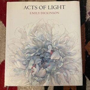 May include: A square book titled Acts of Light by Emily Dickinson. The white cover features a large, detailed watercolor illustration of a blooming peony flower in shades of soft blue, grey, and white with subtle hints of red in the center.