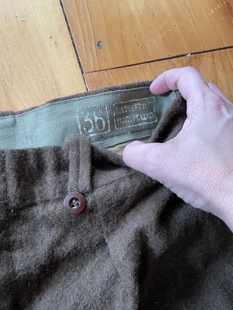 Peut inclure: Un pantalon en laine marron avec un bouton marron et une &eacute;tiquette indiquant "100% EXTRA FINE MERINO WOOL" et "5B".