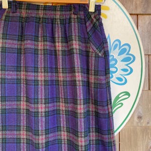 May include: A high-waisted skirt featuring a purple, black, and magenta plaid pattern. The skirt has an elasticized waistband with belt loops and two side pockets. The fabric has a textured appearance and hangs in a straight silhouette.