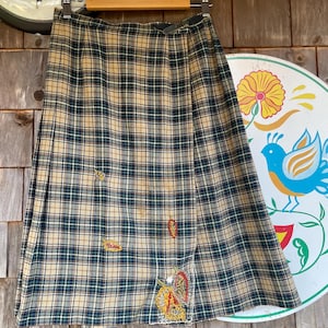 May include: A vintage plaid skirt in beige, navy, and green, hanging on a wooden hanger. The skirt features embroidered patches of leaves and a floral design. The skirt is a classic style, perfect for a retro or vintage-inspired look.