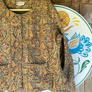 May include: A quilted jacket featuring a mustard yellow, blue, and red paisley floral pattern. The jacket has a round neckline, long sleeves, two front pockets, and a vertical row of silver-toned metal buttons. A label reads Raiment Fashions.