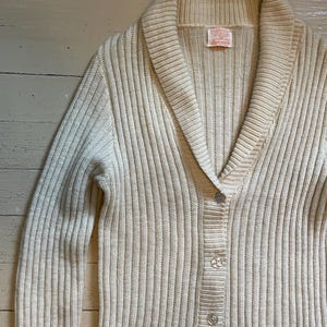May include: A cream-colored cardigan with a shawl collar, ribbed knit, and button closure. The cardigan has two pockets and a label that reads "Pendleton Woolen Mills" with the text "Made in USA" underneath.