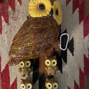 May include: A collection of ceramic owl figurines. The large owl has a yellow head and brown body. Four smaller owls are below. A silver carabiner clip is next to the large owl. The figurines are on a patterned textile.