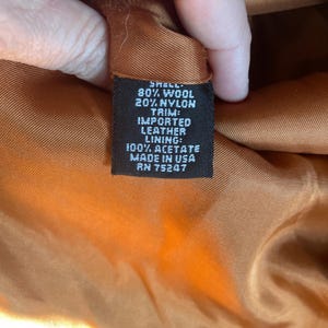 May include: A black fabric label with white text detailing the composition of the garment. The label reads: "Shell: 80% Wool 20% Nylon Trim: Imported Leather Lining: 100% Acetate Made in USA RN 75247"