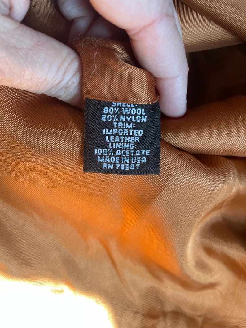 May include: A black fabric label with white text detailing the composition of the garment. The label reads: "Shell: 80% Wool 20% Nylon Trim: Imported Leather Lining: 100% Acetate Made in USA RN 75247"