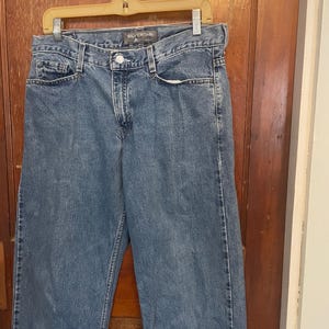 May include: Light blue denim jeans with a button closure and pockets. The jeans are hanging on a hanger. The brand name "SILVERTAB" is visible on the waistband.