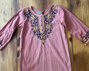 Peut inclure: Une tunique corail à manches longues, ornée de broderies florales noires et jaunes détaillées autour de l'encolure et des poignets. La tunique est faite d'un tissu léger. La broderie est une caractéristique clé.