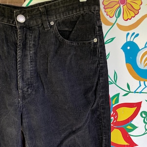 Könnte beinhalten: Nahaufnahme einer schwarzen Cordhose, die an einem Metallbügel hängt. Die Hose hat einen Knopfverschluss, Vordertaschen und ein klassisches Fünf-Taschen-Design. Der Cordstoff hat eine gerippte Textur. Die Hose ist dunkel und einfarbig.