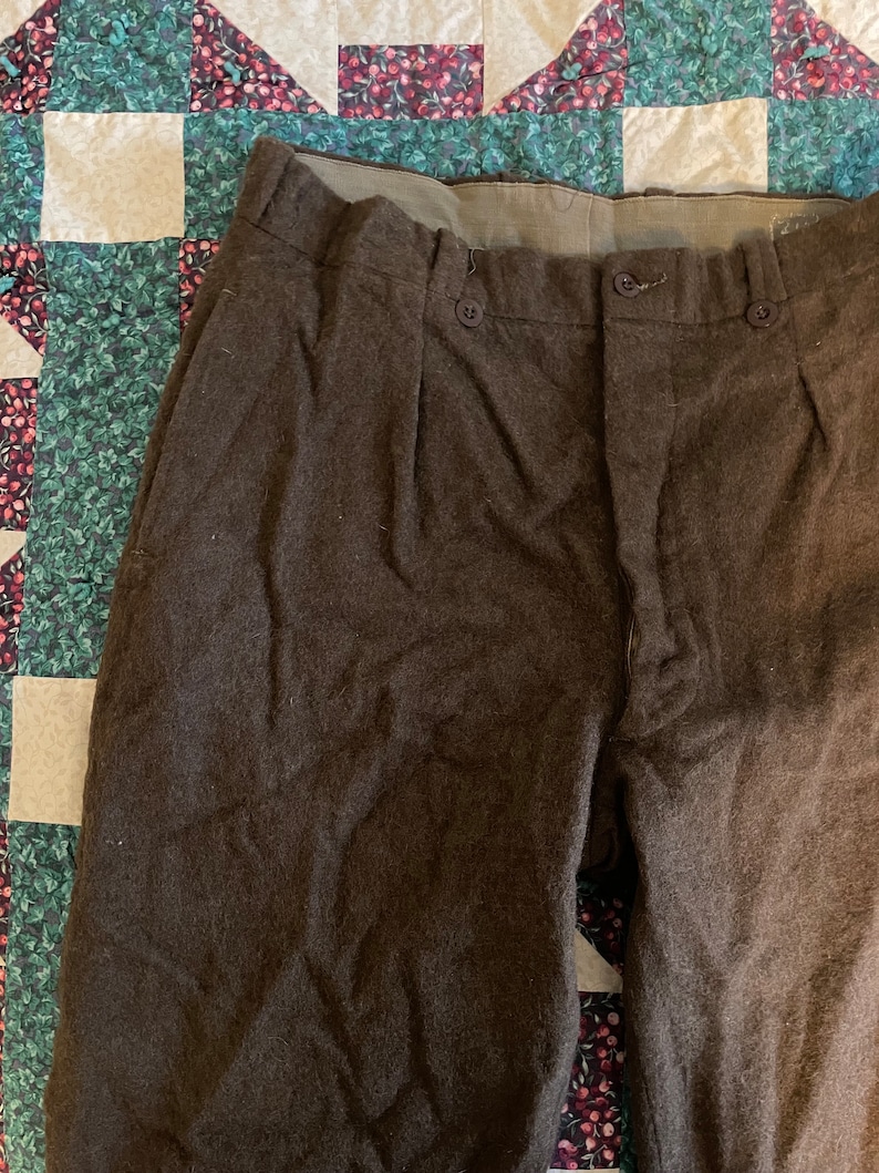 Peut inclure: Un pantalon en laine marron avec une braguette &agrave; boutons et des plis. Le pantalon a une ceinture marron et est fait d'un tissu &eacute;pais et textur&eacute;.