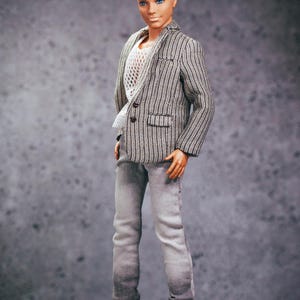 May include: A doll wearing a gray and white striped blazer, white mesh shirt, gray jeans, and white sneakers. The doll has blond hair and blue eyes. The outfit is a modern, casual style.