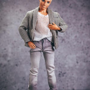 May include: A doll wearing a gray and white striped blazer over a white mesh top, gray jeans, and white sneakers. The doll has blonde hair and blue eyes. The jeans have a faded effect at the bottom.