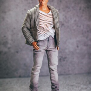 May include: A doll wearing a gray and white striped blazer over a white mesh shirt, gray jeans, and white sneakers. The doll has blonde hair and blue eyes. The outfit is casual and stylish.