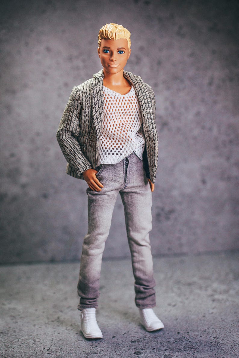 May include: A doll wearing a gray and white striped blazer over a white mesh shirt, gray jeans, and white sneakers. The doll has blonde hair and blue eyes. The outfit is casual and stylish.