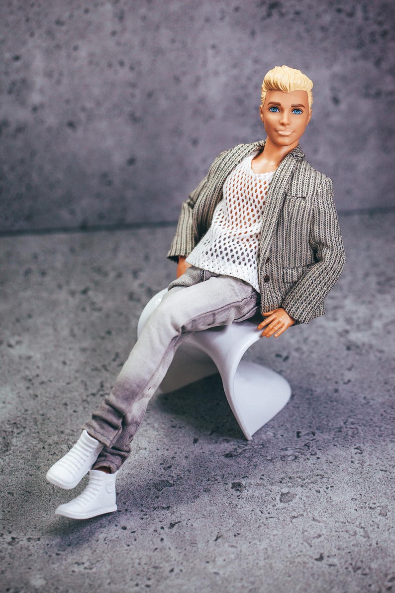 May include: A doll wearing a gray and white striped blazer, a white mesh shirt, and gray pants. The doll is seated on a white chair and wearing white sneakers. The doll has blonde hair and blue eyes.