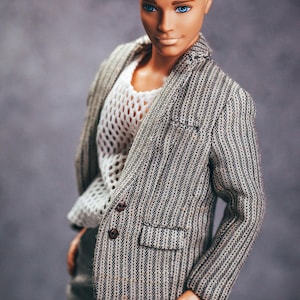 May include: A doll with blonde hair and blue eyes wearing a gray and white striped blazer over a white, open-weave shirt. The doll is also wearing gray pants. The blazer has brown buttons.