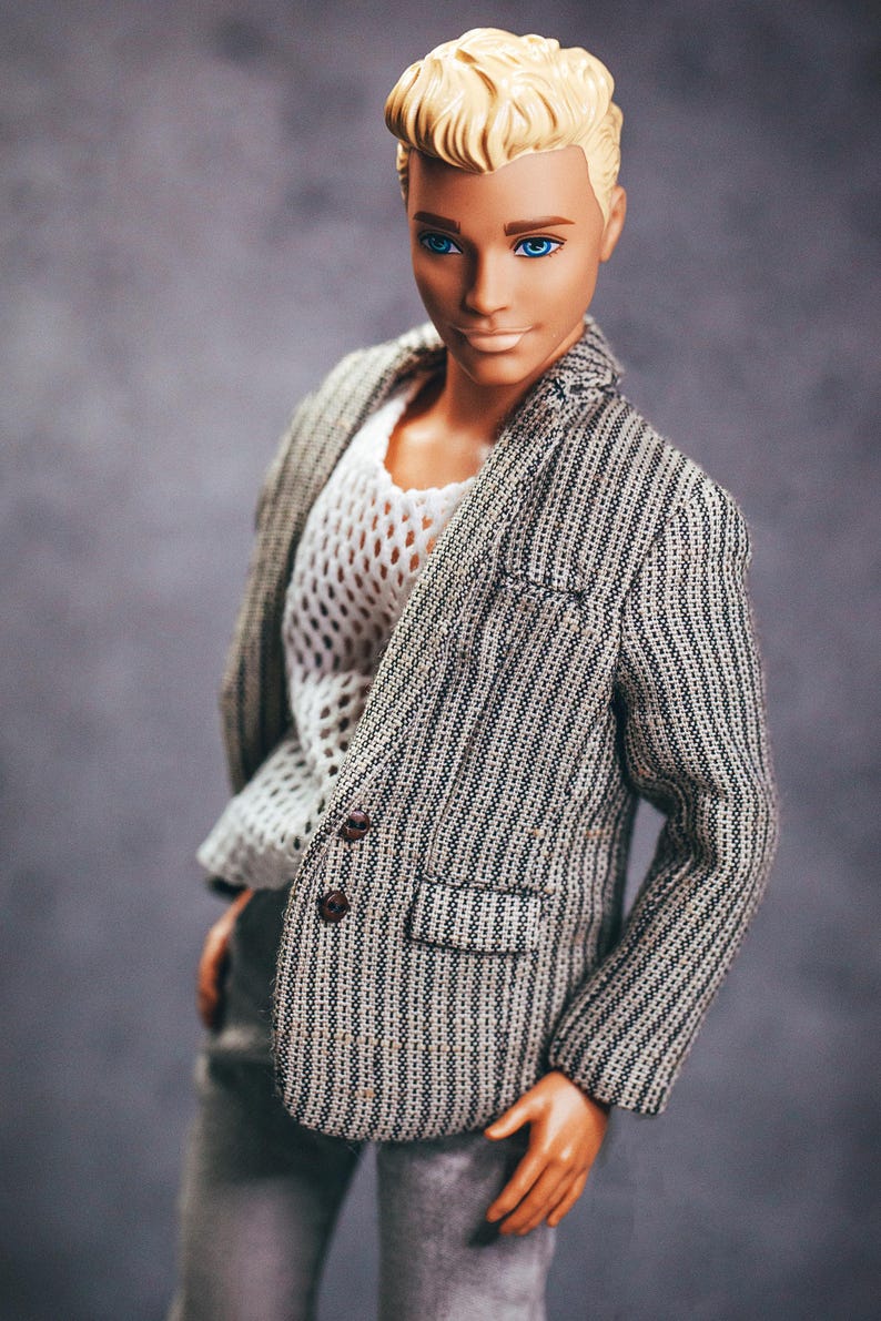 May include: A doll with blonde hair and blue eyes wearing a gray and white striped blazer over a white, open-weave shirt. The doll is also wearing gray pants. The blazer has brown buttons.