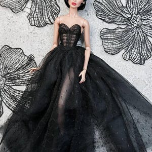 Op de afbeelding: Een modepop in een strapless zwarte jurk met een doorschijnende rok. Het lijfje is gedetailleerd met kant en de pop heeft donker haar en rode lippenstift. Zwarte kanten bloemdecoraties zijn op de achtergrond.