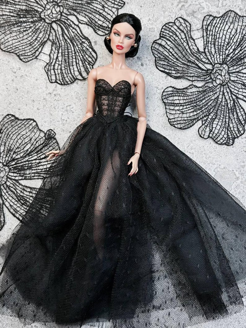 Op de afbeelding: Een modepop in een strapless zwarte jurk met een doorschijnende rok. Het lijfje is gedetailleerd met kant en de pop heeft donker haar en rode lippenstift. Zwarte kanten bloemdecoraties zijn op de achtergrond.