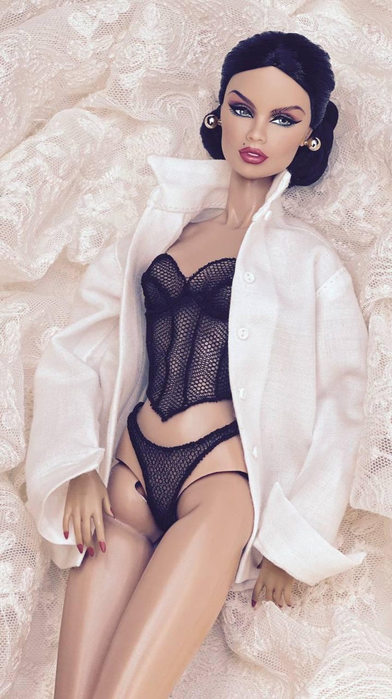 Op de afbeelding: Een pop met een zwart kanten corset en bijpassend ondergoed, met een witte jas met kraag. De pop heeft donker haar, make-up en gouden oorbellen. De pop ligt op een witte kanten stof.