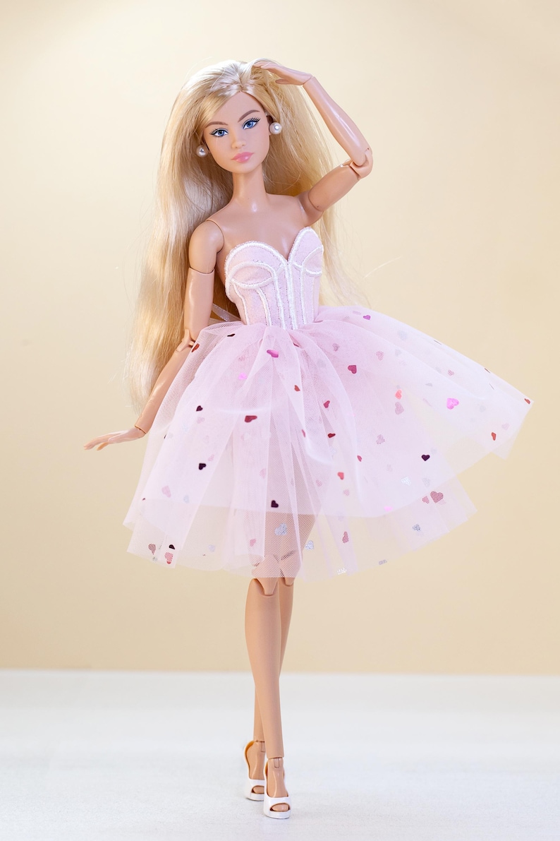 Op de afbeelding: Een modepop met lang blond haar, blauwe ogen en pareloorbellen. Ze draagt een roze, strapless jurk met een tule rok versierd met hartvormige pailletten. De pop poseert met &eacute;&eacute;n arm omhoog, met witte hakken.