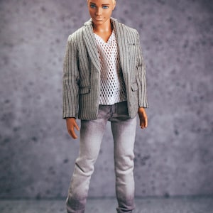 May include: A doll wearing a gray and white striped blazer, a white mesh shirt, and gray jeans. The doll has short, blond hair and is wearing white sneakers. The background is a mottled gray.