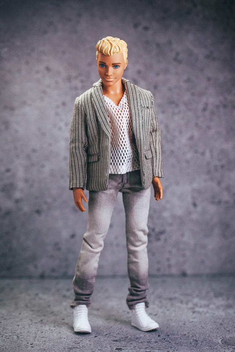 May include: A doll wearing a gray and white striped blazer, a white mesh shirt, and gray jeans. The doll has short, blond hair and is wearing white sneakers. The background is a mottled gray.