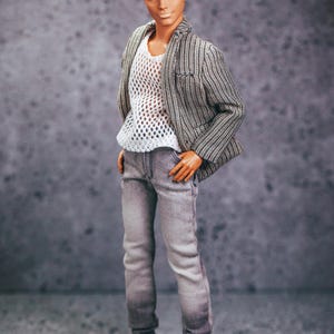 May include: A doll wearing a gray and white striped blazer over a white mesh shirt, gray jeans, and white sneakers. The doll has blond hair and blue eyes. The doll is standing with hands in pockets.