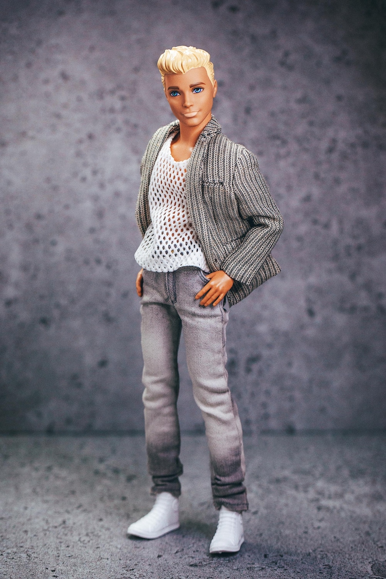 May include: A doll wearing a gray and white striped blazer over a white mesh shirt, gray jeans, and white sneakers. The doll has blond hair and blue eyes. The doll is standing with hands in pockets.