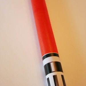 May include: A red and white lightsaber toy with black details. The lightsaber is made of cardboard and has a foil tip.