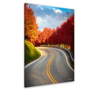 Könnte beinhalten: Eine kurvenreiche Straße mit gelben Doppellinien führt durch eine Landschaft mit leuchtend roten und orangefarbenen Herbstbäumen. Links befindet sich ein grüner Grashügel und darüber ein blauer Himmel mit Wolken.