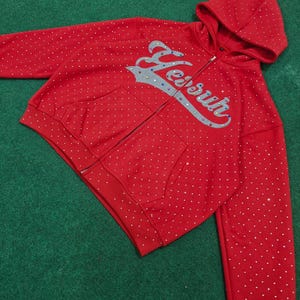K&ouml;nnte beinhalten: Ein leuchtend roter Kapuzenpullover mit Rei&szlig;verschluss und einem durchgehenden Muster aus kleinen wei&szlig;en Punkten. Auf der Vorderseite befindet sich der Schriftzug "Yesur" in einer gro&szlig;en, silberfarbenen Schreibschrift mit bunten Strasssteinen.