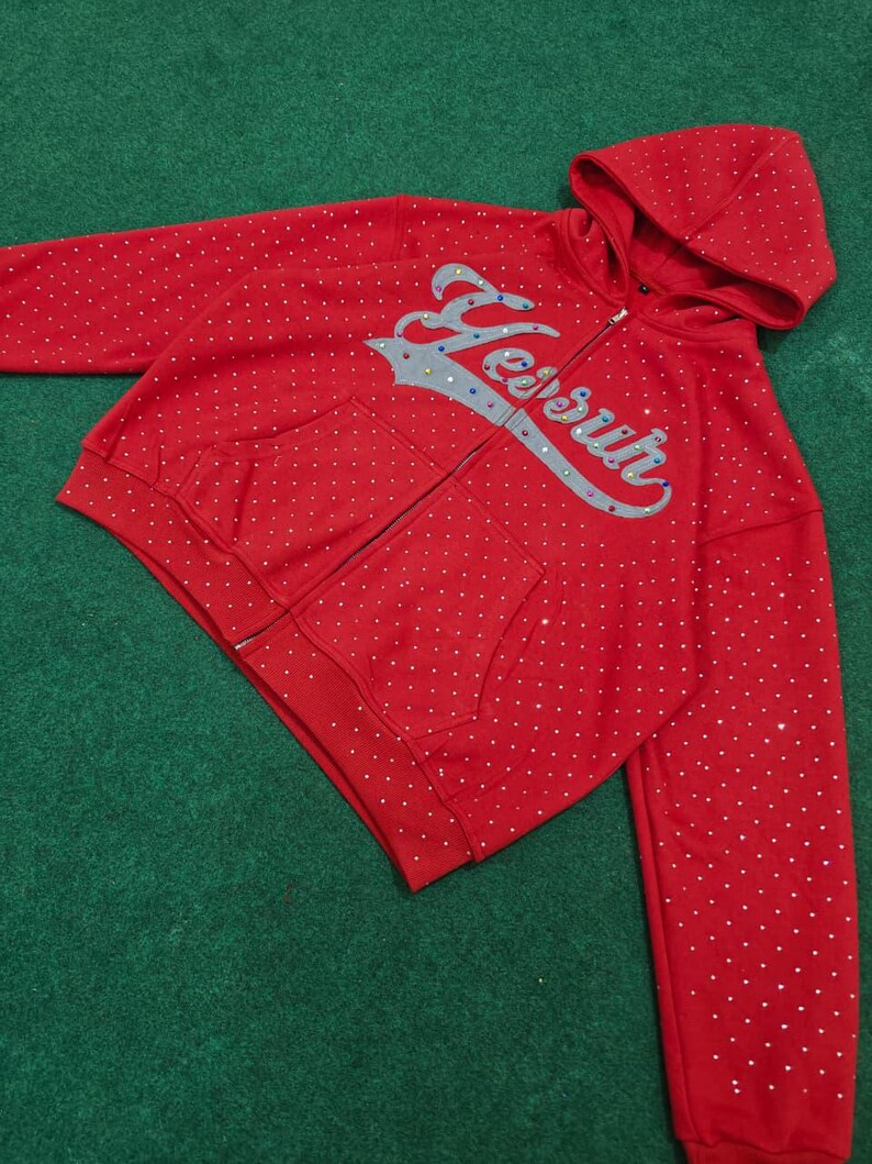 K&ouml;nnte beinhalten: Ein leuchtend roter Kapuzenpullover mit Rei&szlig;verschluss und einem durchgehenden Muster aus kleinen wei&szlig;en Punkten. Auf der Vorderseite befindet sich der Schriftzug "Yesur" in einer gro&szlig;en, silberfarbenen Schreibschrift mit bunten Strasssteinen.