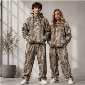Puede incluir: Un conjunto de dos piezas a juego compuesto por una sudadera con capucha y pantalones holgados, ambos con un estampado de camuflaje en tonos marrón, gris y bronceado, con zapatillas blancas en los pies.