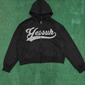 K&ouml;nnte beinhalten: Ein schwarzer Kapuzenpullover mit Rei&szlig;verschluss und langen &Auml;rmeln, verziert mit einem Muster aus kleinen wei&szlig;en Nieten. Auf der Vorderseite steht das Wort Yessuh in einer gro&szlig;en, stilisierten wei&szlig;en Schreibschrift mit bunten Nieten.
