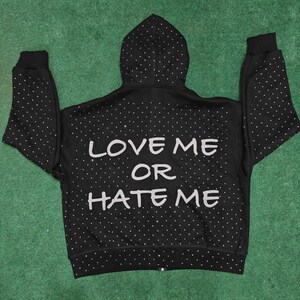 K&ouml;nnte beinhalten: Ein schwarzer Kapuzenpullover mit Rei&szlig;verschluss und einem durchgehenden Muster aus kleinen wei&szlig;en Punkten. Auf der R&uuml;ckseite befindet sich der Text "LOVE ME OR HATE ME" in gro&szlig;en, hellen Gro&szlig;buchstaben. Das Kleidungsst&uuml;ck hat eine Kapuze.