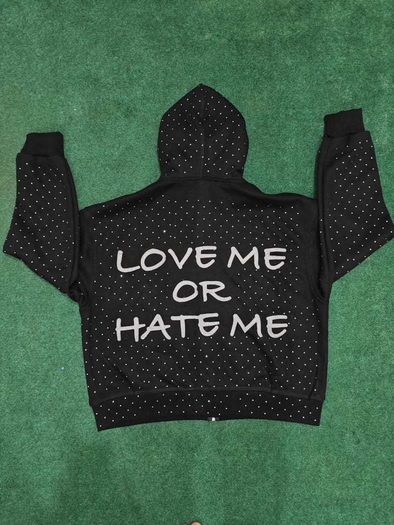 K&ouml;nnte beinhalten: Ein schwarzer Kapuzenpullover mit Rei&szlig;verschluss und einem durchgehenden Muster aus kleinen wei&szlig;en Punkten. Auf der R&uuml;ckseite befindet sich der Text "LOVE ME OR HATE ME" in gro&szlig;en, hellen Gro&szlig;buchstaben. Das Kleidungsst&uuml;ck hat eine Kapuze.