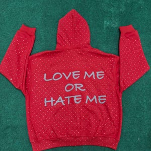 K&ouml;nnte beinhalten: Ein leuchtend roter Kapuzenpullover mit einem verstreuten Muster aus kleinen silbernen Strasssteinen auf dem gesamten R&uuml;cken, den &Auml;rmeln und der Kapuze. Der Text "LOVE ME OR HATE ME" ist in einer gro&szlig;en, hellgrauen, stilisierten Schrift aufgedruckt.