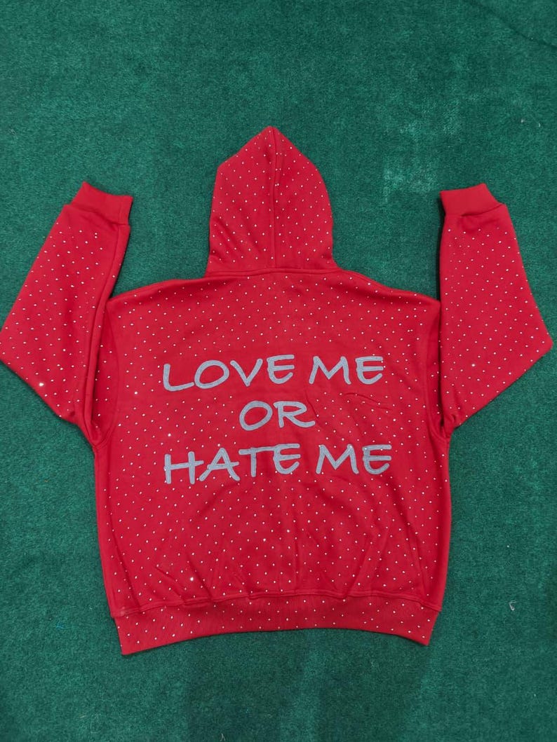 K&ouml;nnte beinhalten: Ein leuchtend roter Kapuzenpullover mit einem verstreuten Muster aus kleinen silbernen Strasssteinen auf dem gesamten R&uuml;cken, den &Auml;rmeln und der Kapuze. Der Text "LOVE ME OR HATE ME" ist in einer gro&szlig;en, hellgrauen, stilisierten Schrift aufgedruckt.