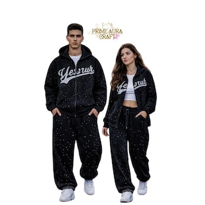 Puede incluir: Un conjunto de sudadera negra con cremallera y pantalones de chándal a juego, con un patrón de motas blancas y el texto "Yessir" en letra cursiva en el pecho. La sudadera incluye una capucha y una cremallera frontal para el cierre.