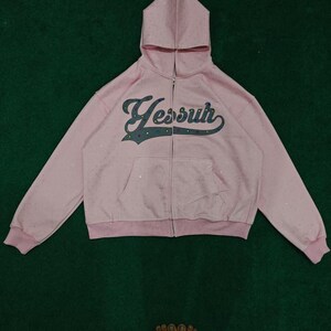 K&ouml;nnte beinhalten: Ein hellrosa Kapuzenpullover mit Rei&szlig;verschluss, Kapuze, langen &Auml;rmeln und einer K&auml;ngurutasche vorne. Auf der Brust befindet sich der Schriftzug Yessuh in dunkelblauer Schreibschrift mit kleinen, bunten Strasssteinen auf dem Stoff.