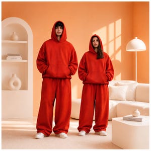 May include: Two individuals wearing matching bright red hooded sweatshirts and loose-fitting red sweatpants. Each hoodie features a large front kangaroo pocket and a drawstring-free hood. The garments are solid red with a plain, uniform texture.