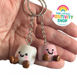 May include: Two marshmallow-shaped keychains, one white and one pink, each with a smiling face and brown feet. The keychains are attached to silver key rings and chains. The Little Positivity Shop logo is visible in the background.