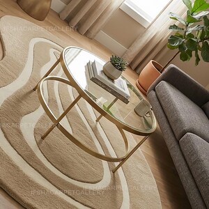 Op de afbeelding: Een moderne woonkamer met een gouden glazen salontafel, boeken en een kleine plant in een pot. Onder de tafel ligt een beige en wit patroonkleed en rechts een grijze bank.