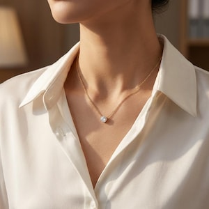 May include: A gold necklace with a single, round, clear gemstone pendant. The necklace is worn against a white collared shirt. The pendant is the focal point, catching the light and creating a sparkling effect.