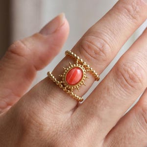 May include: Gold-toned ring featuring a coral-colored oval stone with a sunburst design. The ring has a double band with beaded detailing. The ring is a piece of jewelry.