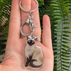 Può includere: Un portachiavi con gatto siamese con catena e chiusura color argento. Il gatto ha occhi blu, corpo beige e segni marrone scuro. Il portachiavi è tenuto in mano su uno sfondo di fogliame verde.