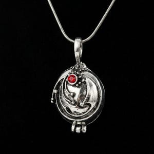 May include: A silver-toned metal locket pendant suspended from a matching thin snake chain. The circular locket features an antiqued finish with raised swirling lines and a single small, round, faceted red gemstone set near the top center.